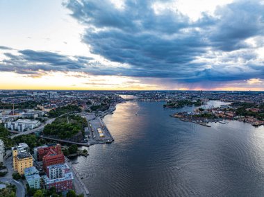 Stockholm 'ün çarpıcı günbatımı manzarası, Saltsjon sularında yelkenli tekneleriyle birlikte, dramatik bulutlarla çerçevelenmiş ve kentsel manzarayı İsveç' in doğasıyla harmanlayan Curgarden dahil..