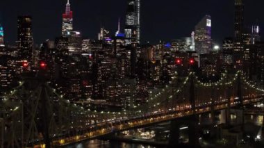 Hava görüntüleri gece New York şehrinin ufuk çizgisini yakalıyor, Queensboro köprüsünü ve East River 'ı gözler önüne seriyor, Manhattan silueti arka planında kentsel bir sahne oluşturuyor..