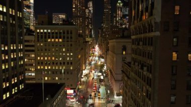 Arabalar, gece vakti Manhattan, New York 'ta aydınlık gökdelenlerle çevrili hareketli bir caddede yol alırlar. Şehir manzarasının canlı enerjisi ve modern mimarisi.