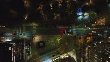 Arabalar, otobüsler ve sarı taksiler gece vakti Manhattan, New York 'ta yoğun bir kavşakta yol alırlar. Yayalar kaldırımlarda ve yaya geçitlerinde yürür, gökyüzünden bakarlar..