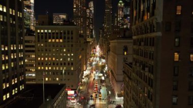 Arabalar, gece vakti Manhattan, New York 'ta aydınlık gökdelenlerle çevrili hareketli bir caddede yol alırlar. Şehir manzarasının canlı enerjisi ve modern mimarisi.