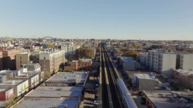 New York 'un Queens semtindeki yerleşim yerlerini gezen yükseltilmiş tren gökyüzü perspektifinden aydınlık güneş ışığı altındaki binalar ve caddelerle şehir manzarasını gözler önüne seriyor..