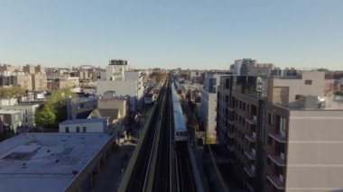 Yükseltilmiş metro treni Queens 'teki meskun New York bölgesinde hareket ediyor. Gün ışığında metropoliten gökdeleni görünen apartmanların önünden geçiyor..