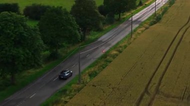 Havadan bakıldığında, Belçika 'da bir kır yolunda ilerleyen siyah bir araba, yemyeşil tarlalardan ve büyüleyici evlerden geçiyor, kırsal manzaranın dingin güzelliğini vurguluyor..