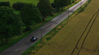 Araba Belçika 'da bir köy yolunda gidiyor. Çevresindeki manzara yeşil ve yemyeşil, ekin ve ağaç tarlaları var..