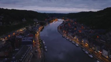 Aydınlatılmış binalar, Belçika 'nın Dinant kentindeki Meuse nehrinin kıyısında uzanır. Alacakaranlık, büyüleyici şehrin üzerine dingin bir ışık saçar ve güzel bir akşam manzarası yaratır..
