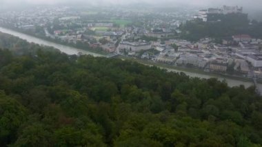 Salzburg, Avusturya 'nın hava manzarasında sisli bir günde Salzach Nehri, ağaçlar, binalar ve Hohensalzburg Kalesi var..