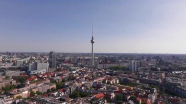 Hava görüntülerinde Alexanderplatz 'daki ikonik Berlin TV kulesi, sıcak güneş ışığıyla aydınlatılarak engin bir şehrin üzerinde yükseliyor. Geniş binalar ve sokaklar her yöne doğru uzanıyor..