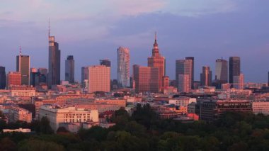 Varşova şehir merkezinin modern gökdelenleri batan güneşin sıcacık ışığında yıkanıyor. Finansal bölgenin hemen dışındaki bir şehir parkını çevreleyen yeşil ağaçlar.