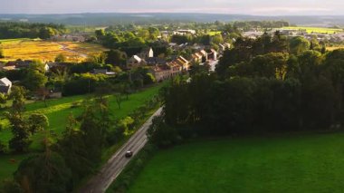 Belçika kırsalındaki küçük bir köye giden yol boyunca giden siyah bir arabanın hava görüntüsü. Güneş parlıyor ve gökyüzü biraz bulutla mavi..
