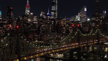 Hava görüntüleri gece New York şehrinin ufuk çizgisini yakalıyor, Queensboro köprüsünü ve East River 'ı gözler önüne seriyor, Manhattan silueti arka planında kentsel bir sahne oluşturuyor..