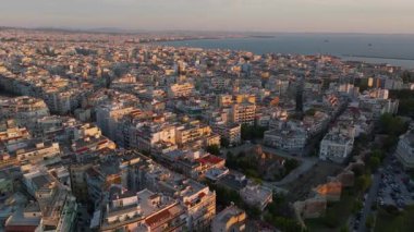 Selanik, Yunanistan, gün batımının ılık renklerinde parlıyor. Binalar ve sokaklar Ege Denizi 'ne yükseltilmiş bir perspektiften uzanırken şehir hayatı gelişiyor..