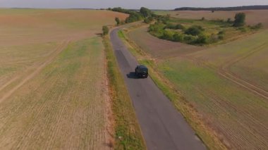 Hava görüntüleri, kırsal yaz yolculuklarının cazibesini gözler önüne seren, canlı tarım alanlarıyla çevrili, dolambaçlı bir kır yolu boyunca seyahat eden siyah bir SUV yakalıyor..