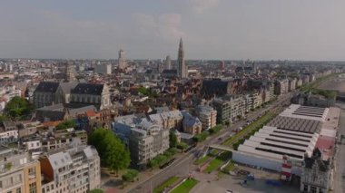 Antwerps canlı şehir merkezinde kalabalık caddelerde hareket eden arabalar ve tramvaylar, ikonik mimari ve arka planda çarpıcı bir katedral kulesi yer alıyor..