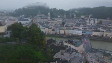 Salzburg, Avusturya şehir merkezinde Salzach Nehri 'nin şehri sardığı hava manzaralı sergiler. Hohensalzburg Kalesi arka planda yükselir, sisle kaplanır. Salzburg, Avusturya 5 Temmuz 2024