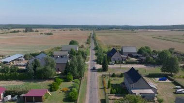 Havadan bakıldığında düz bir yolda seyreden parlak siyah bir araba görülüyor. Canlı tarlalar ve bir köydeki antika evler, çarpıcı bir kırsal kesim manzarası yakalıyor..