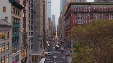 Arabalar ve otobüsler, Madison Square Park yakınlarındaki kalabalık bir Beşinci Cadde boyunca ilerliyorlar. Flatiron Bölgesi 'nin yüksek binaları tarafından kuşatılmışlar ve alacakaranlıkta yüksek bir noktadan görülmüşler..