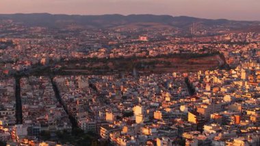 Selanik, Yunanistan, batan güneşin altın parıltısıyla güneşleniyor. Şehir manzarası aşağılara kadar uzanıyor, şehir yoğunluğu ve yeşil alan ceplerinin karışımını ortaya çıkarıyor..
