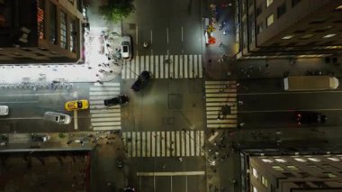 Gece vakti Manhattan, New York 'ta, eşsiz bir hava perspektifinden işlek bir kavşaktan geçen sarı taksiler ve arabalar tripod çerçeveleri çerçeveler. Yayalar caddeyi geçiyor.