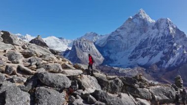 Dağcı, kırmızı ceketli, duacı bayrakları dalgalanan kayalık yamaçlarda, Gus 'ın arkasında yükselen Everest tepelerinde gezinir..