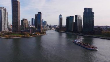 Hava görüntüleri, Gotham Point, Long Island City, Queens gökdelenleri ve yüksek gökdelenleri tarafından çerçevelenen Doğu Nehri boyunca hareket eden bir mavnayı çekiyor..