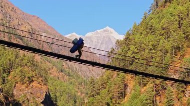Mevsimlik dağ rehberi Himalaya vadisinin yukarısındaki tehlikeli asma köprüyü geçiyor. Karla kaplı Everest Dağı engebeli alp panoramasına hakim..