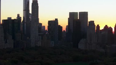 Central Park 'tan izlenen Manhattan şehir merkezi silueti üzerinde günbatımında sıcak bir parıltı var. Sahne, gündüz akşam dönüşümleriyle şehir hayatının güzelliğini yakalıyor..