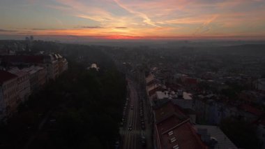 Arabalar Prag 'da ağaçlarla kaplı bir sokakta dolaşırken güneş batıyor, şehir manzarasını ılık ışıkta yıkıyor ve canlı bir alacakaranlık ortamı yaratıyor..