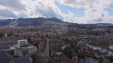Bern şehir manzarasını gözler önüne seren hava manzarası, Aare Nehri, İsviçre 'nin bulutlu gökyüzünün altındaki Gurten Dağı' nın yanında dönüyor..