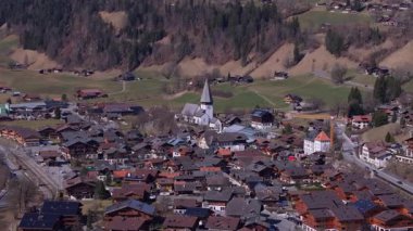 Hava görüntüsü, İsviçre Alp manzarasında yuva yapmış Gstaad köyünü, yemyeşil dağ vadisini, geleneksel chalets ve St Mauritius Kilisesini gözler önüne seriyor..
