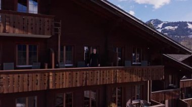 İsviçre Alpleri manzarasına bakan balkonda yürüyen genç bir adam ve dağ zirvelerinin arasına kurulmuş Gstaad köyü..