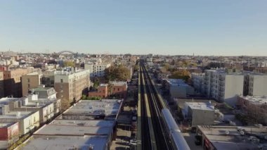 New York 'un Queens semtindeki yerleşim yerlerini gezen yükseltilmiş tren gökyüzü perspektifinden aydınlık güneş ışığı altındaki binalar ve caddelerle şehir manzarasını gözler önüne seriyor..