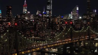 Hava görüntüleri gece New York şehrinin ufuk çizgisini yakalıyor, Queensboro köprüsünü ve East River 'ı gözler önüne seriyor, Manhattan silueti arka planında kentsel bir sahne oluşturuyor..