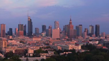 Havadan bakıldığında Varşova ufuk çizgisi, yaz günbatımında çarpıcı bir mor gökyüzüne karşı canlı renklerle aydınlatılmış modern binalar içeriyor..