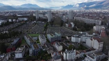Hava aracı perspektifi genişleyen Grenoble Saint Roch mezarlığını, İle Verte konutunu, gri kış bulutlarının altında yükselen Alp zirvelerini gözler önüne seriyor..