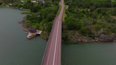 Finlandiya, Aland 'da deniz üzerinde bir köprüden geçen beyaz bir arabanın kuş bakışı görüntüsü. Sahne, açık mavi gökyüzünün altındaki çarpıcı takımadaları yakalıyor..