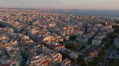 Selanik, Yunanistan, gün batımının ılık renklerinde parlıyor. Binalar ve sokaklar Ege Denizi 'ne yükseltilmiş bir perspektiften uzanırken şehir hayatı gelişiyor..
