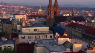 Prag çatıları ve kilise kuleleri güneşin altın parıltısında güneşleniyor. Şehir panoraması aşağıda sergileniyor, büyüleyici mimari cazibesini sergiliyor..