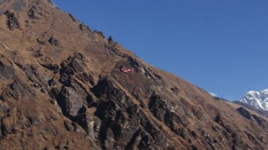 Nepal 'in uzak dağlık bir bölgesinin havadan görünüşü, engebeli arazi üzerinde uçan kırmızı bir helikopter ve karla kaplı zirveler..