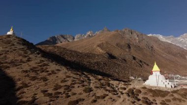 Nepal dağlarına kurulmuş sakin bir köyün havadan görünüşü, sarı tepeli geleneksel beyaz stupalar ve arka planda görkemli karlı zirveler.
