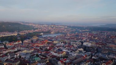 Güneş ışığı Prag şehir merkezindeki binaları aydınlatır, ortasından akan bir nehir ve uzaktaki tepeler.