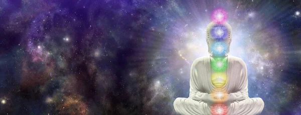 Meditating Chakra Buddha sitting in lotus position surrounded by  deep space - buddha on right side with seven chakras against a starry dark blue celestial sky with a massive nebula and copy space for text