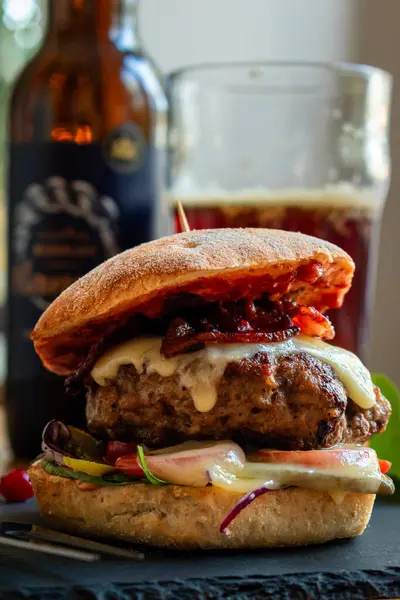 Homemade bacon cheeseburger on a dark tile with berries on the side and cold beer in the background. Food porn and nasty burger, smash burger