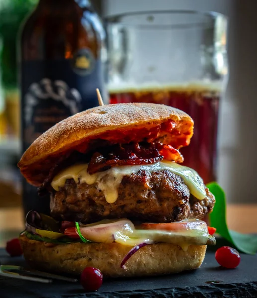 Homemade bacon cheeseburger on a dark tile with berries on the side and cold beer in the background. Food porn and nasty burger, smash burger
