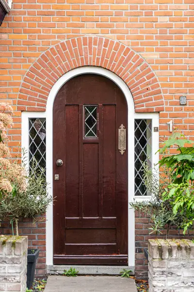 Wooden front door of a home. Front view of a wooden front door on a brick house with windows plants and a wide view of the porch and front walkway. Horizontal shot.
