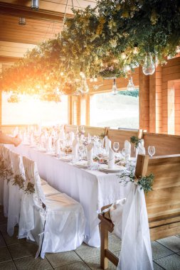 Büyük, uzun, süslü, ahşap bir masa örtüsü ve tabaklar, çiçekler, mumlar ile kaplı sandalyeler doğada ormanın yakınında duruyor. Düğün ziyafeti. ışık arkaplanı