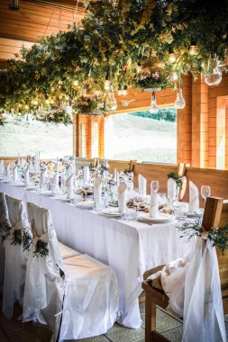 Büyük, uzun, süslü, ahşap bir masa örtüsü ve tabaklar, çiçekler, mumlar ile kaplı sandalyeler doğada ormanın yakınında duruyor. Düğün ziyafeti. ışık arkaplanı