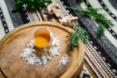 Yuvarlak kesme tahtasındaki un içinde kırılmış yumurta, peçeteli koyu ahşap masa, üst manzara.