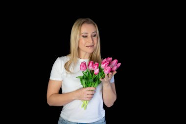 Beyaz tişörtlü mutlu sarışın kadın siyah arka planda dururken elinde bir sürü lale tutuyor.