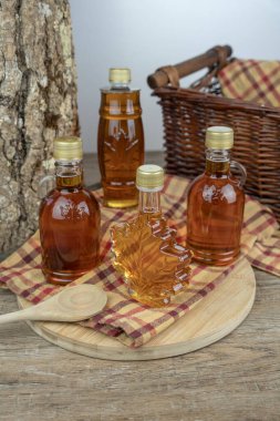 Bottles of maple syrup displayed on a wooden table with cutting board, wooden spoon, basket and checkered tablecloth.  Delicious.  New.  Maple Syrup production.  Maple sugaring. Alternative sweetener. Amber.  Golden. 